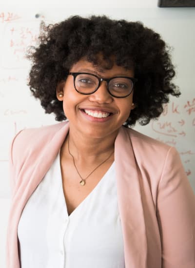 A younger, black female in white top and pink blazer smiling with whiteboard behind her